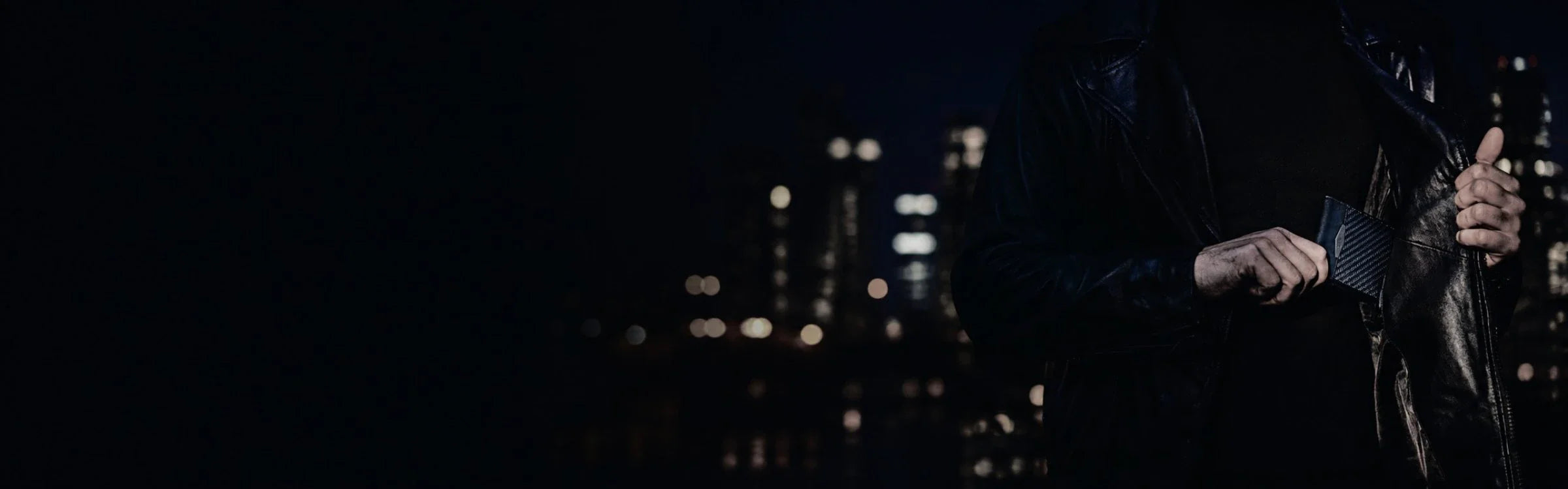 Man in leather jacket pulling a carbon fiber COLDFIRE wallet from inner pocket against urban night skyline
